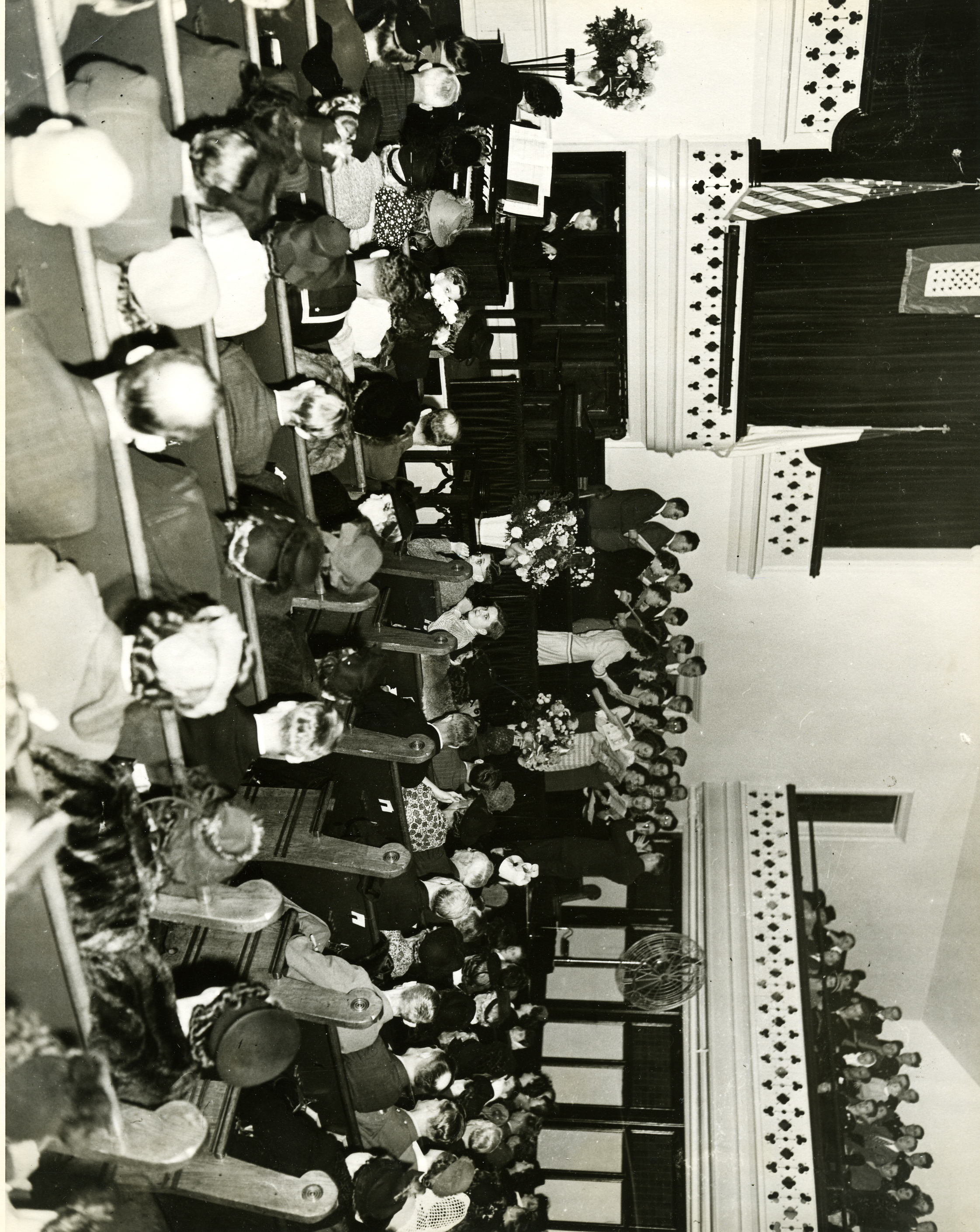A black and white photo of a crowded church. People are seated in pews, and a choir is standing and singing on stage.
