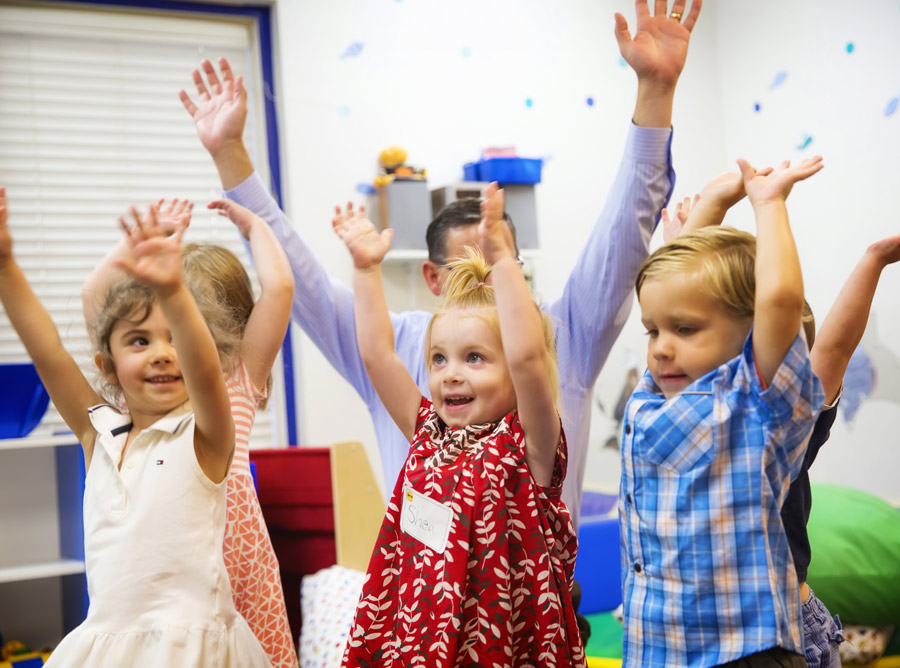 A group of four young children raise their arms enthusiastically in a lively classroom.