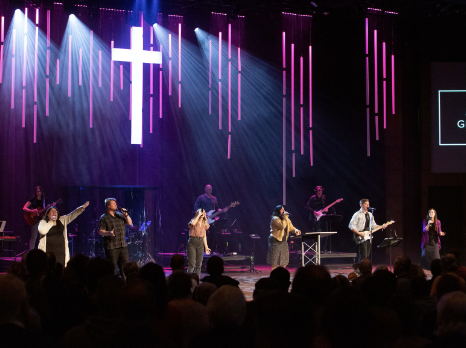 A band composed of musicians and singers lead worship on a stage with purple lights and a cross in the background.